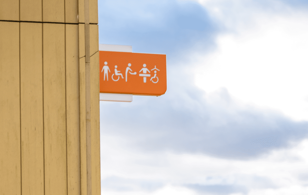 Image: Orange sign with toilet signs, onside the side of a building with a blue and white sky in the background.