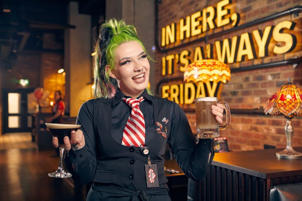 IMAGE: A person with green hair holding two drinks in a brown restaurant. 