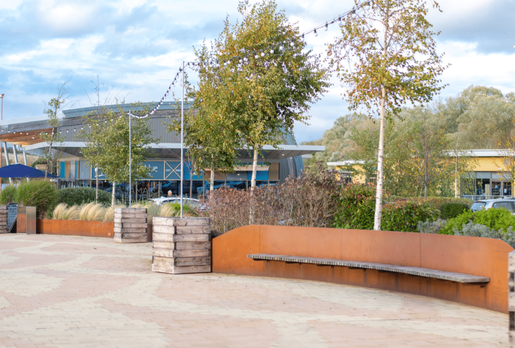 Image: Outside of Rushden Lakes, with a wooden bench and trees in the background.