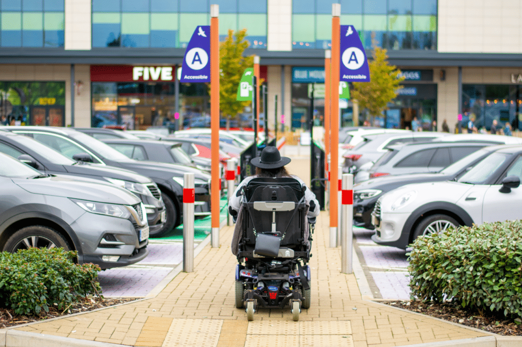 Image: Back of a wheelchair, with a disabled women sitting in it, wearing a black hat. Background shows two Accessible Bays, and the front of stores.