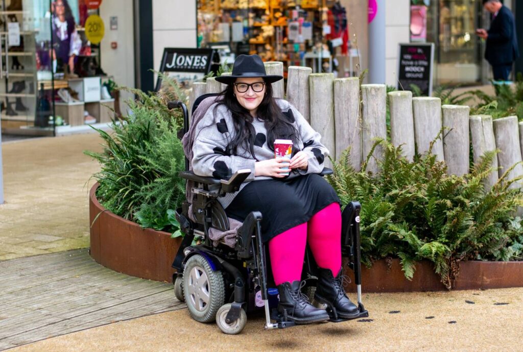 Image: Disabled woman sat in a wheelchair, in the Garden Square area of Rushden Lakes, wearing grey jumper with black hearts, pink rights, black skirt and black hat.